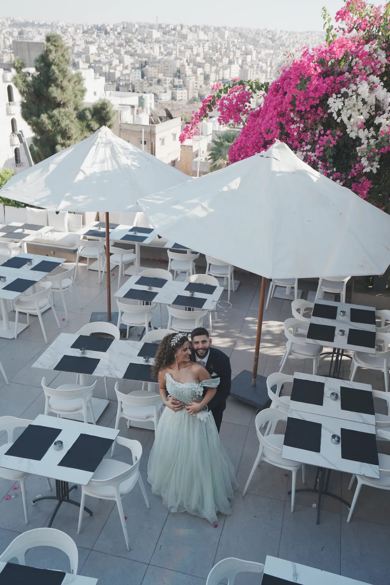 A couple embracing on a rooftop restaurant terrace, seen from above between white umbrellas, with Amman and bougainvillea beyond