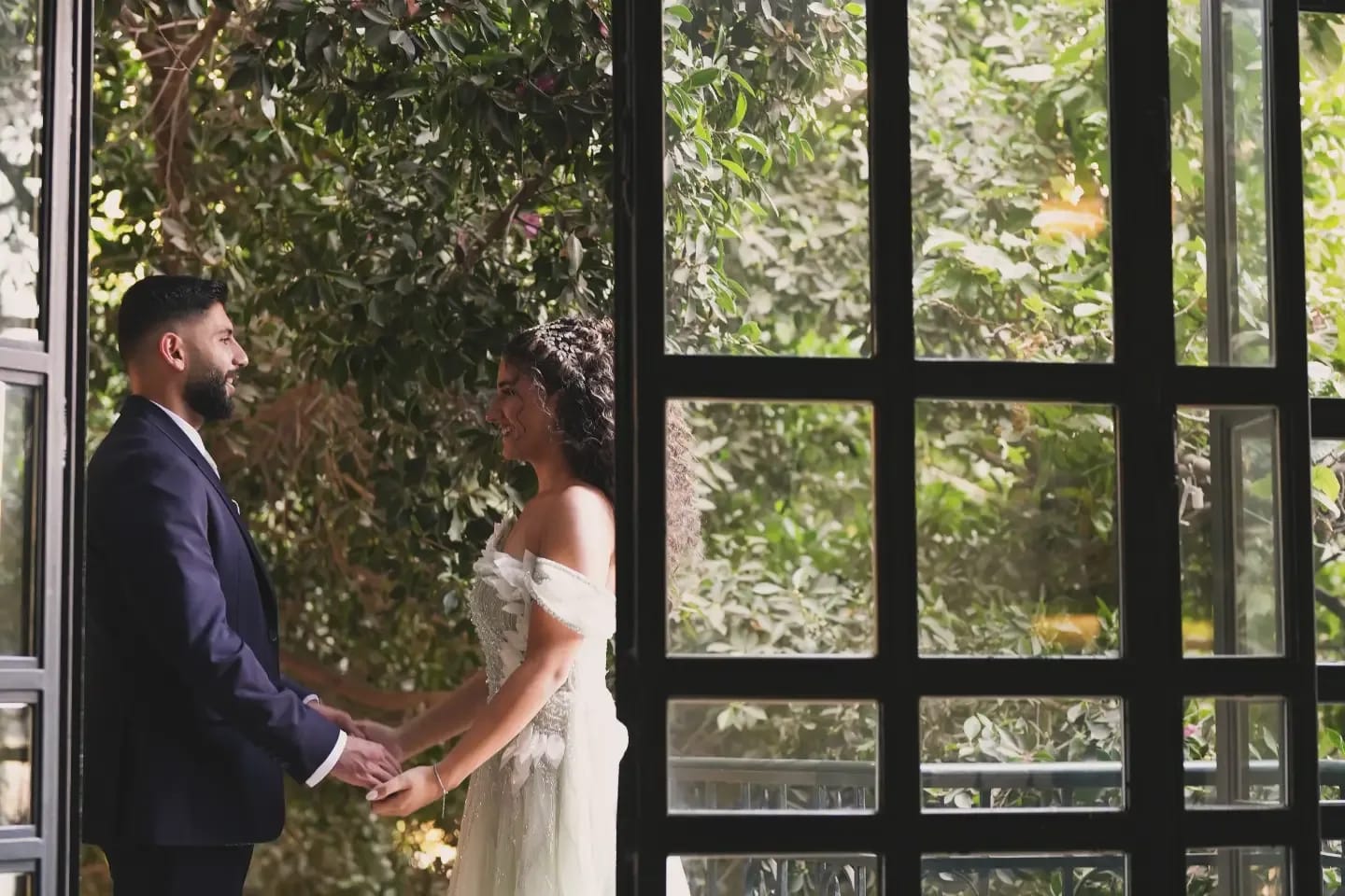 A couple holding hands, framed by a dark window with green leaves behind
