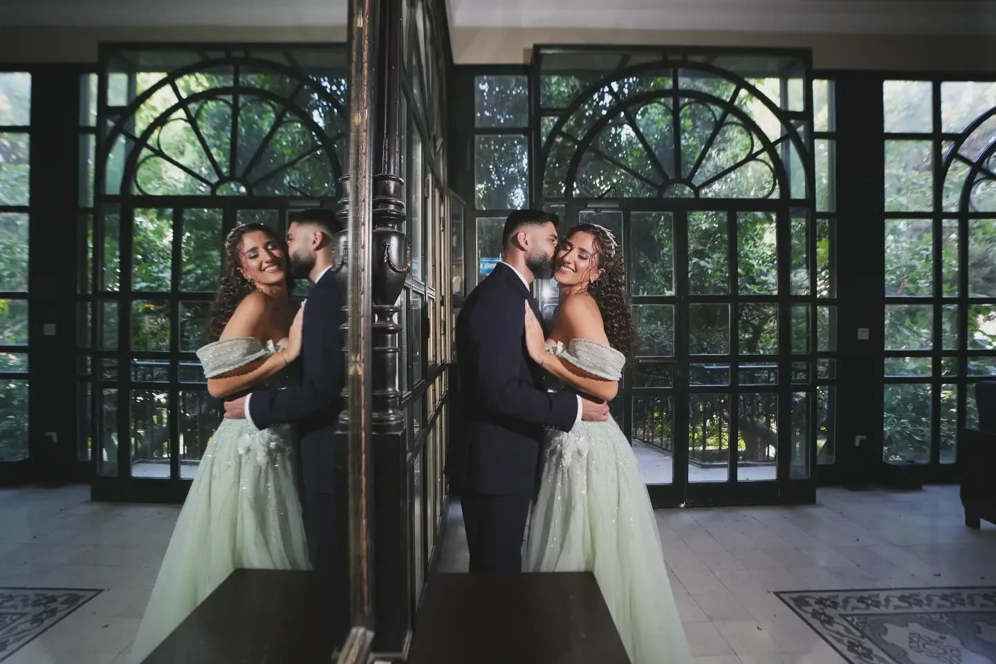 A groom kissing his bride's cheek, mirrored in a tall dark-framed mirror