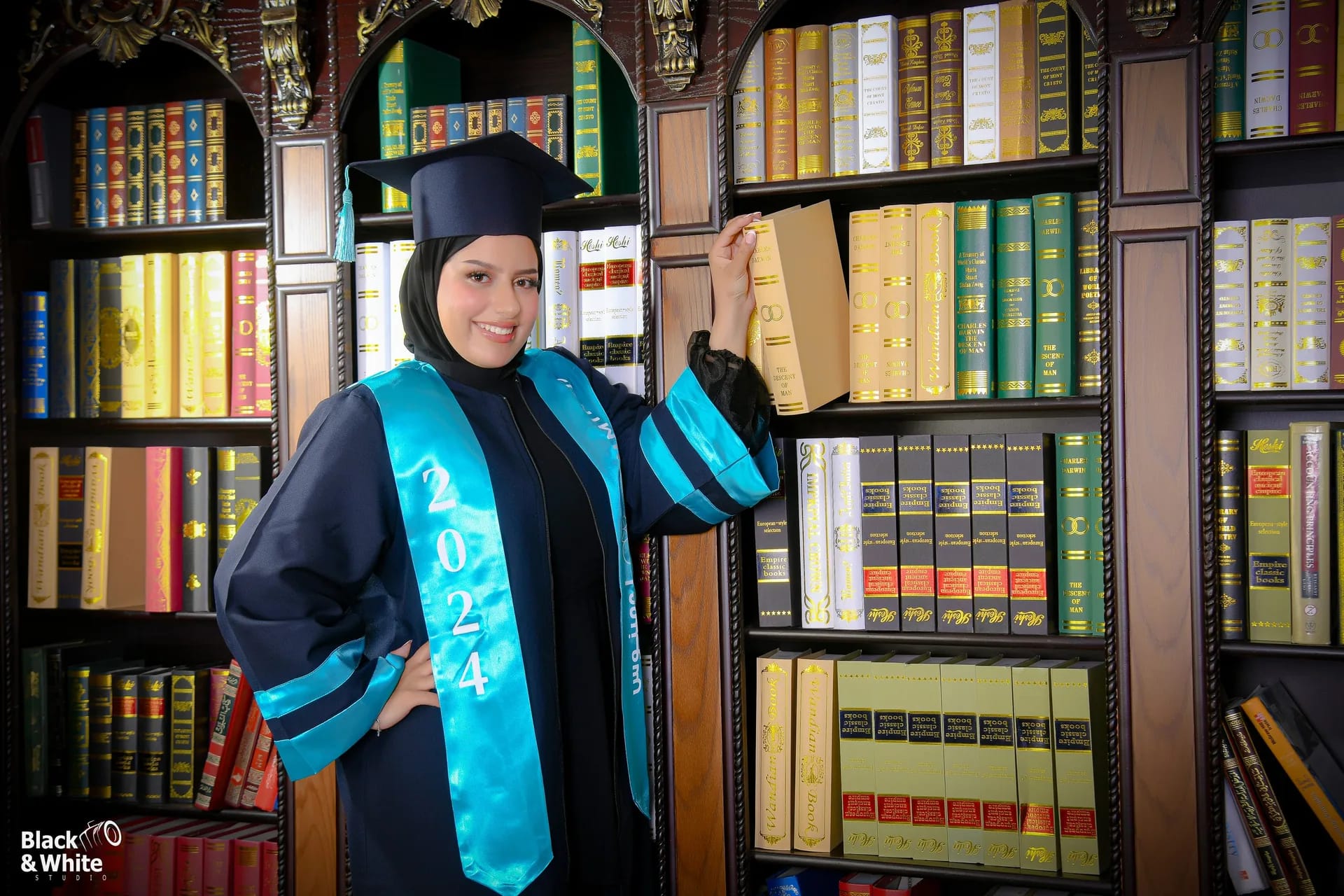 A graduate in a navy gown and teal sash beside the bookshelves