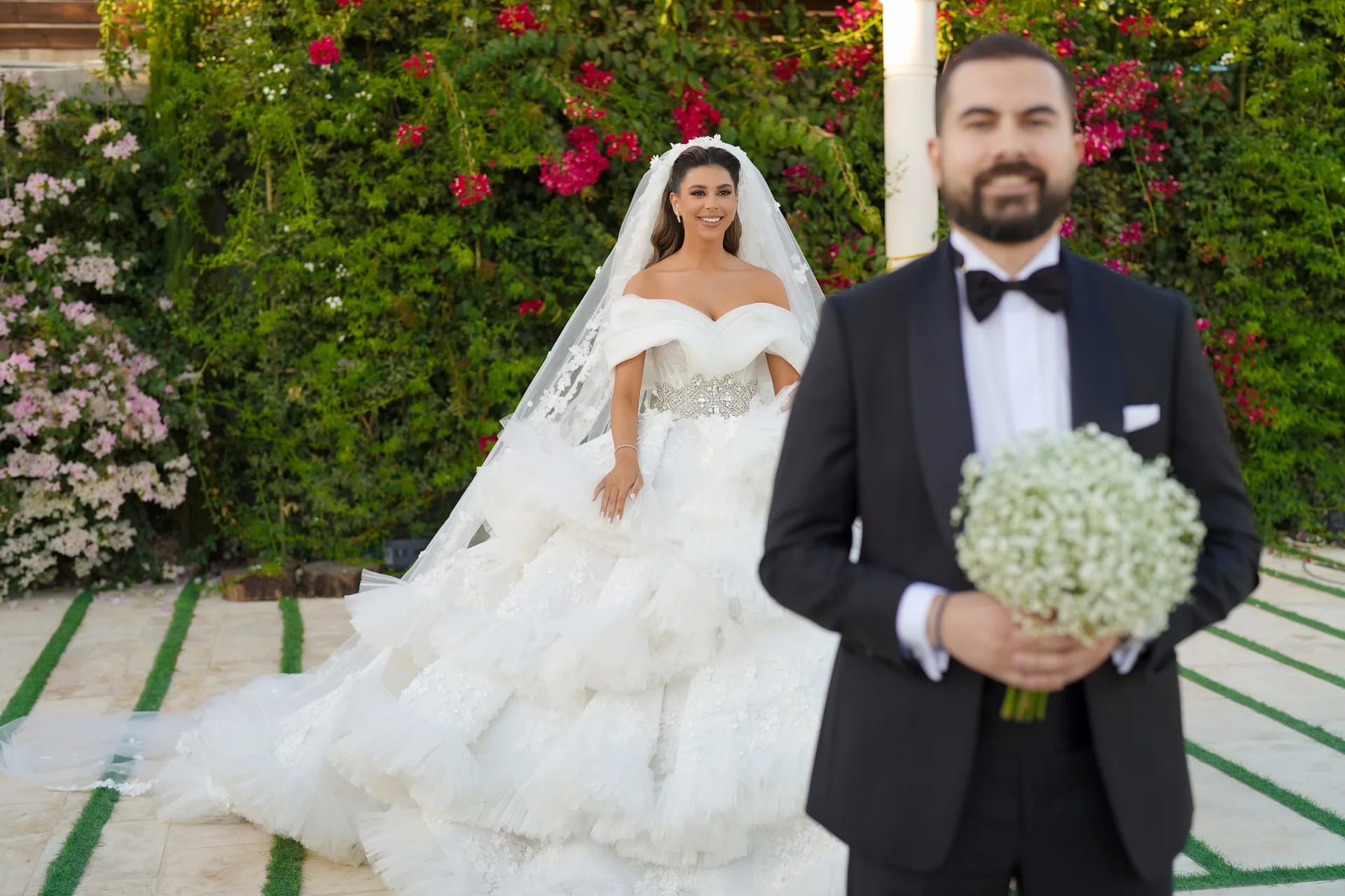 A groom holding a bouquet beside his bride in front of a wall of roses