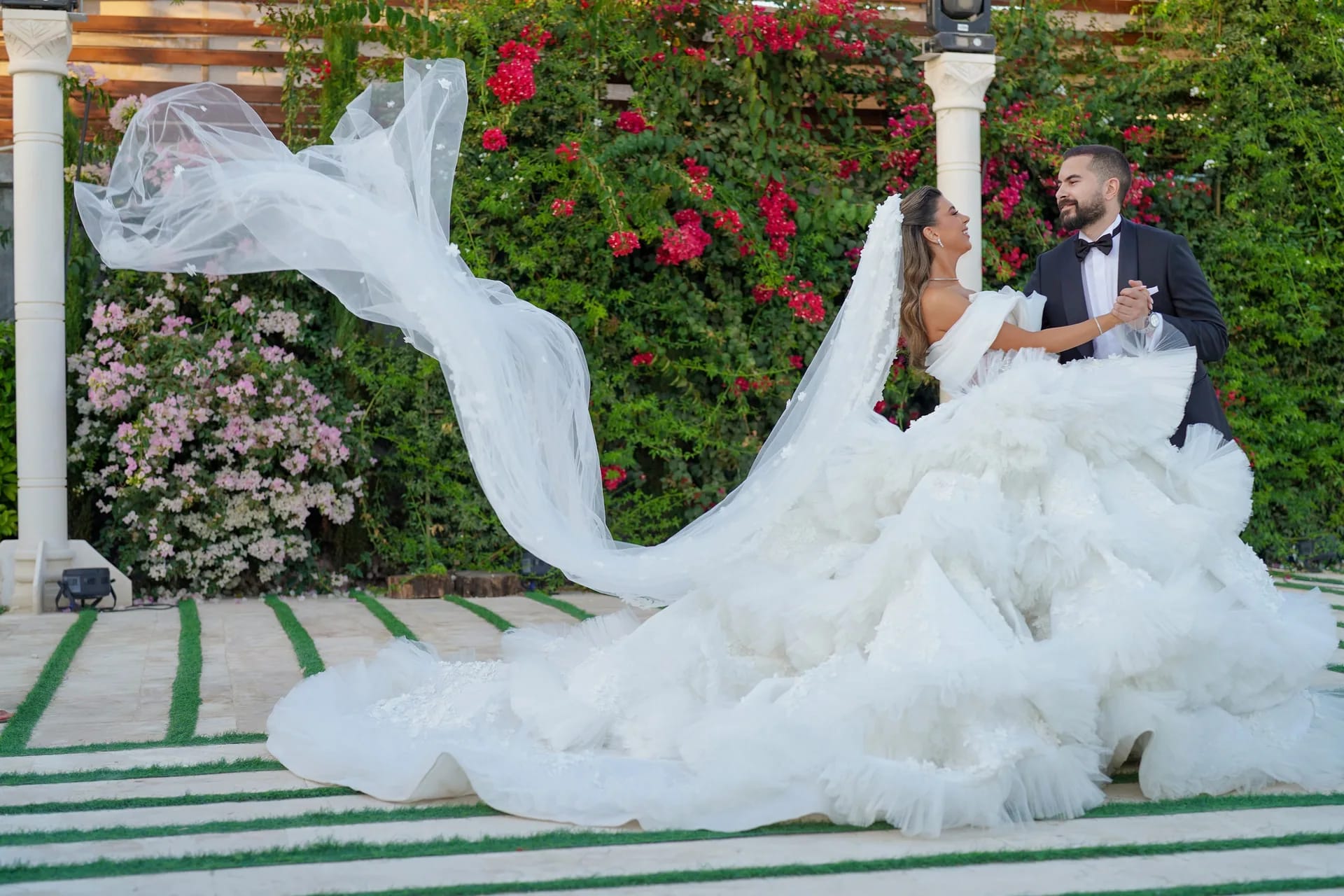 A bride's train sweeping across the lawn beside a wall of flowers