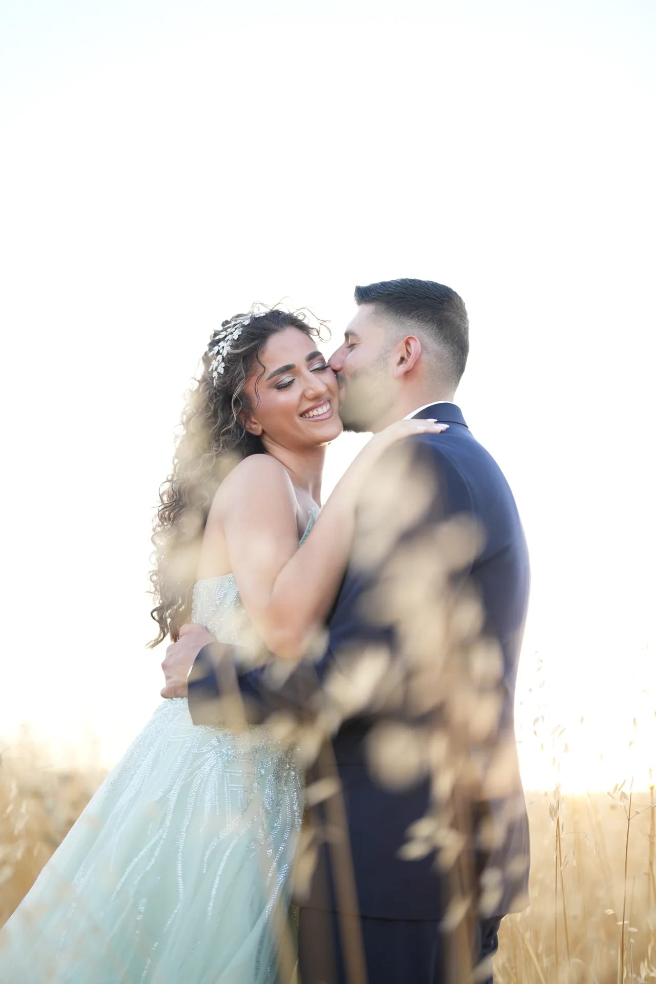 A groom kissing his bride's cheek in a golden field at sunset, grasses blurred in front