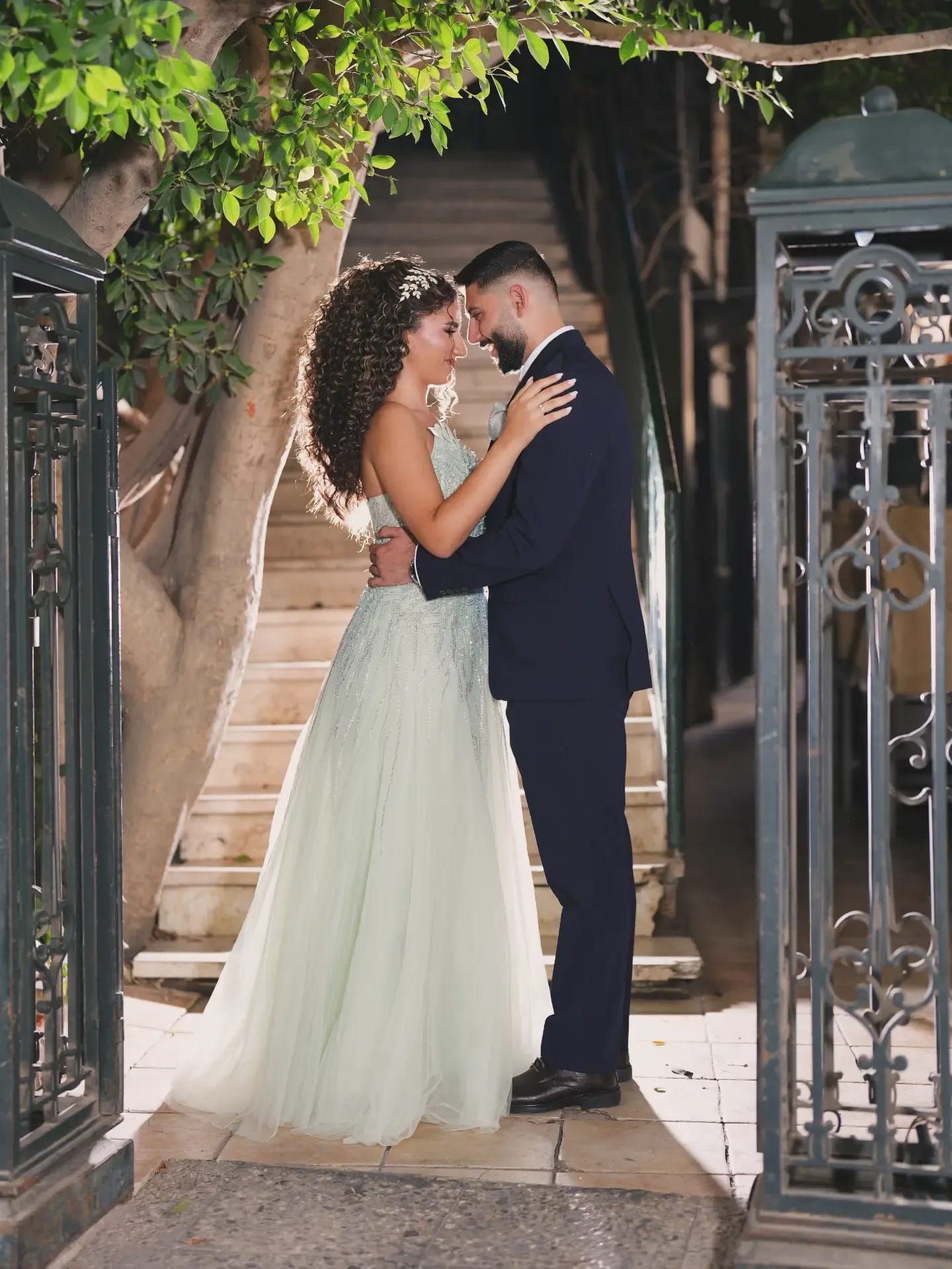 A couple embracing forehead to forehead between two iron gates, a staircase behind them