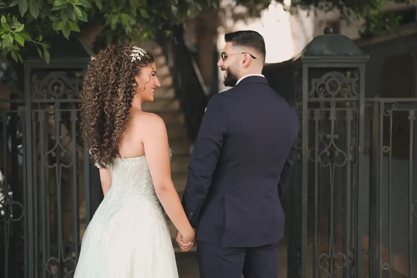 A couple holding hands, glancing at each other as they walk toward an old iron gate