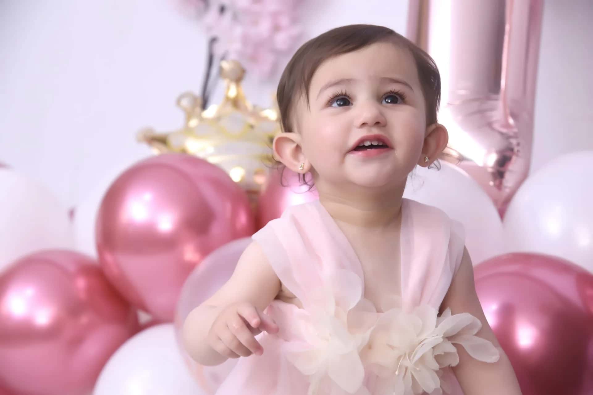 A baby in a pink dress looking up among balloons