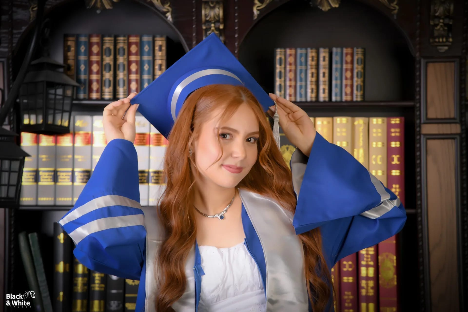 A graduate in a blue gown adjusting her cap in front of a library set