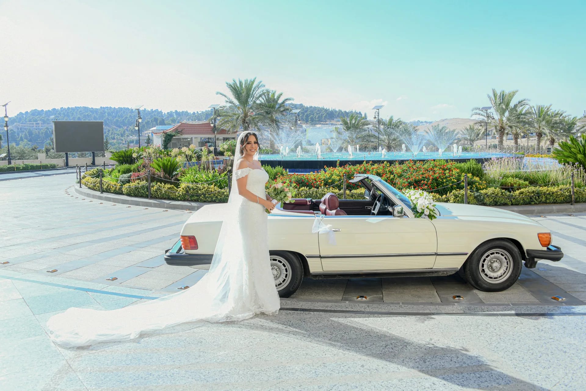 A bride beside a classic white car on a sunny avenue of palms